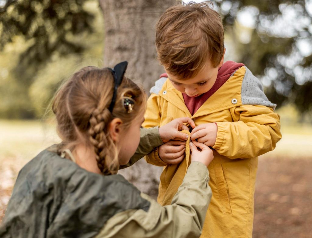 Момиче показва жест на доброта, помагайки на по-малко дете да си закопчее якето.